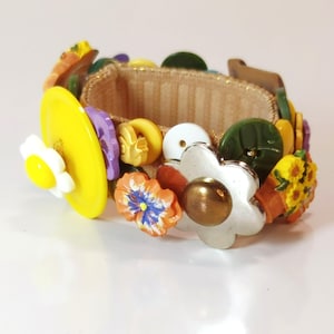 May include: A colorful button bracelet featuring a variety of shapes and colors. The bracelet includes yellow, white, purple, orange, and green buttons, with flower and leaf designs. The band is a tan fabric.