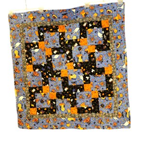 Peut inclure: Un quilt patchwork carré sur le thème d'Halloween avec des blocs alternés de tissu orange, noir et bleu clair. La courtepointe présente des motifs de citrouilles, de fantômes et de chats, entourés d'une fine bordure noire à motifs de toiles d'araignée.