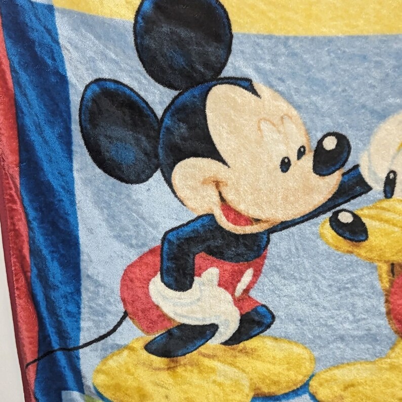 May include: A blue, red, and yellow fleece blanket featuring Mickey Mouse and Pluto. Mickey Mouse is smiling and looking to the right. Pluto is looking to the left.