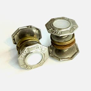 May include: A pair of silver-toned metal cufflinks featuring an octagonal face with a central white circular inlay. The stem of each cufflink consists of stacked brass and copper-colored rings between the octagonal ends.