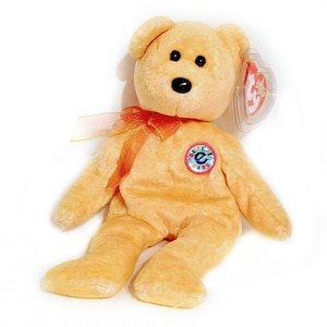 Peut inclure: Un ours en peluche orange clair avec des yeux et un nez noirs, et un petit ruban orange noué autour du cou. L'ours a un écusson circulaire coloré sur la poitrine et une étiquette en forme de cœur attachée à l'oreille.