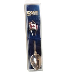 May include: A souvenir set featuring a spoon and a pin with a Canadian flag design. The pin has a red and white flag with a maple leaf. The spoon is silver-colored. The packaging reads "EGMB Souvenirs Made in Canada."