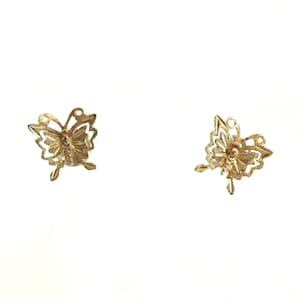 May include: Two gold-colored butterfly charms. Each charm features a detailed, openwork design with delicate wings and a central body. The charms are likely intended for jewelry making or crafting, and are set against a white background.