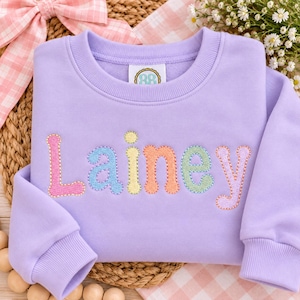 May include: A light purple crewneck sweatshirt featuring the name Lainey embroidered across the chest in multi-colored, bubble-style letters. Each letter is outlined with a decorative stitch, and the interior label displays the text 8B.
