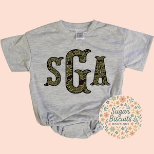 May include: A gray short-sleeved baby onesie with the letters "SGA" in a camouflage pattern. The onesie has a round neckline and snap closures at the bottom. The brand "Simpli Blanks" is printed on the tag. A floral logo for "Sugar Biscuits Boutique" is in the lower right corner.