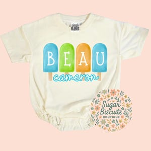 May include: Cream-colored baby bodysuit with a popsicle design spelling "BEAU" in blue, green, and orange. The name "cameron" is written below the popsicles in a script font. The bodysuit has short sleeves and a snap closure.