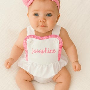 May include: A baby wearing a white romper with a pink gingham trim and the name "Josephine" embroidered in pink. The baby also wears a matching pink gingham bow headband. The outfit is designed for infants.