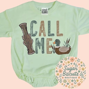 May include: Pale green baby bodysuit with the words "CALL ME" in brown and teal letters. A duck and duck call graphic are featured on the front. The bodysuit has a round neckline and short sleeves.