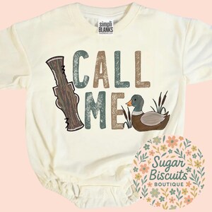 May include: Cream baby bodysuit featuring the words "CALL ME" in brown, teal, and tan lettering. A cartoon duck and a duck call graphic are also featured on the front. The bodysuit has short sleeves and snap closures.