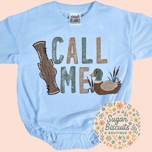 May include: Pale blue baby bodysuit featuring the words "CALL ME" in green, brown, and tan. The design includes a cartoon duck and a brown hunting call. The bodysuit is from Simpli Blanks.