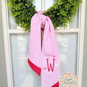May include: Pink and white striped scarf with red trim, embroidered with the letter "W" and small hearts. The scarf is hanging on a green wreath. The scarf is made of a soft, textured fabric.