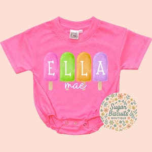 May include: A bright pink baby bodysuit with a popsicle design. The popsicles are in shades of pink, green, orange, and purple, each with a letter spelling "ELLA" and the name "mae" below. The bodysuit has short sleeves and snaps at the bottom.