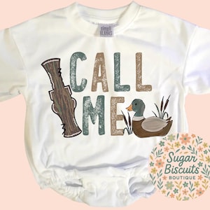 May include: White baby bodysuit with the words "CALL ME" in green, brown, and tan. A cartoon duck and duck call are featured in the design. The Sugar Biscuits Boutique logo is in the lower right corner.