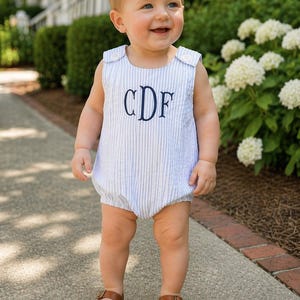 May include: A baby wearing a light blue and white striped romper with the embroidered initials "CDF" in navy blue. The baby is also wearing brown leather sandals with buckles.