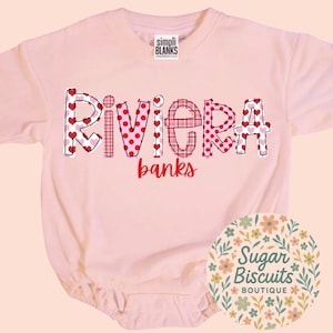 May include: A light pink baby bodysuit with the name "Riviera banks" in a decorative font. The letters are decorated with hearts and plaid patterns. The bodysuit has short sleeves and a round neckline. A circular logo with floral elements is in the bottom right corner.
