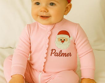 May include: A baby wearing a pink onesie with a Santa Claus graphic and the name "Palmer" embroidered on the front. The baby is also wearing a red bow headband. The baby is sitting on a white surface.