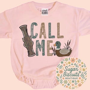 May include: A pale pink baby bodysuit featuring the text "CALL ME" in green and brown. The design includes a duck and a duck call. The bodysuit has short sleeves and a round neckline.
