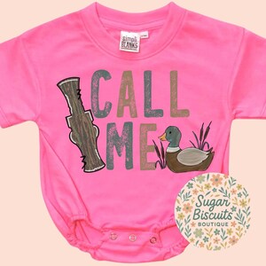 May include: A vibrant pink baby bodysuit featuring the text "CALL ME" in various colours and a duck illustration. The bodysuit has short sleeves and popper fastenings. The label "SIMPLI BLANKS" is visible.