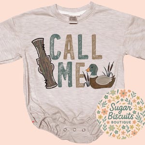 May include: A light grey baby bodysuit with the words "CALL ME" in teal and brown block letters. A cartoon duck and duck call are also featured. The Sugar Biscuits Boutique logo is in the lower right corner.