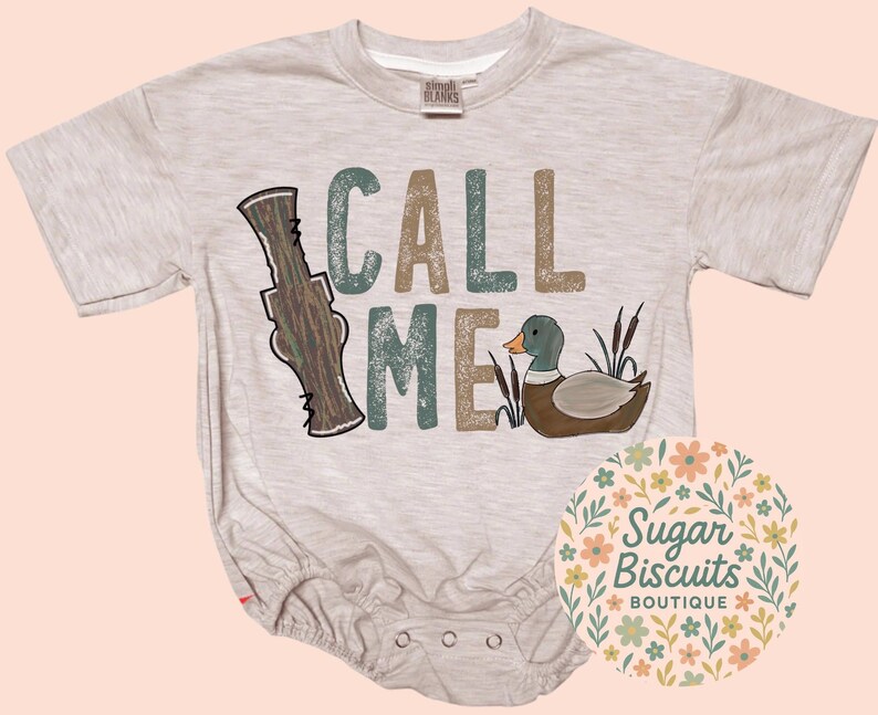 May include: A light grey baby bodysuit with the words "CALL ME" in teal and brown block letters. A cartoon duck and duck call are also featured. The Sugar Biscuits Boutique logo is in the lower right corner.