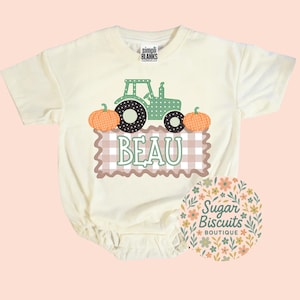 May include: A cream-colored short-sleeve bodysuit featuring a graphic of a green polka-dot tractor flanked by two orange pumpkins. Below the tractor is a brown and white gingham rectangle with the name BEAU printed in light green capital letters.