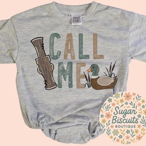 May include: A light grey baby onesie with the words "CALL ME" in green and brown letters. A cartoon duck and duck call are also featured. The onesie has short sleeves and a round neckline. The Sugar Biscuits Boutique logo is in the lower right corner.