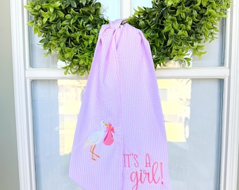May include: A pink and white striped baby shower towel with the embroidered text "IT'S A girl!" and a stork carrying a pink bundle. The towel is hanging from a green wreath.