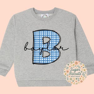 May include: A light gray sweatshirt featuring a large, blue and white gingham "B" with the name "baylor" in a cursive font. The sweatshirt has long sleeves and a round neckline. A floral logo for "Sugar Biscuits Boutique" is on the bottom right.