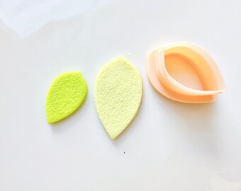 Puede incluir: Dos piezas de fondant en forma de hoja verde y un cortador de galletas en forma de hoja de plástico sobre una superficie blanca.