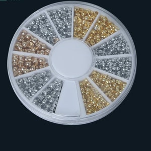 May include: A white plastic wheel with 12 compartments filled with small silver, gold, and rose gold beads. The beads are for nail art and can be used to decorate nails.