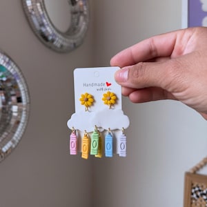 Könnte beinhalten: Handgefertigte Ohrringe mit gelben Gänseblümchen-Ohrsteckern, weißen Wolkenakzenten und bunten Buntstift-Anhängern. Auf der Karte steht "Handmade with love".