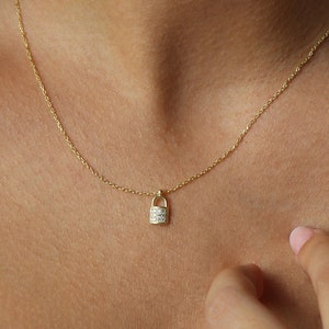 Puede incluir: Un collar de cadena dorada con un pequeño colgante de candado incrustado con piedras de circonita cúbica transparentes.