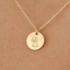 Puede incluir: Un collar dorado con un pequeño colgante redondo. El colgante presenta una ilustración grabada de un perro. El collar se muestra sobre un fondo color piel.