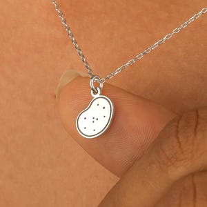 May include: A silver-colored necklace featuring a small, bean-shaped pendant. The pendant has a white outline and small black dots. The necklace chain is delicate and appears to be made of silver. The jewelry is displayed against a skin-toned background.