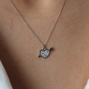 Puede incluir: Un collar de plata con un colgante en forma de corazón con una pequeña flecha que lo atraviesa. El corazón está cubierto de pequeñas piedras de circonita cúbica transparentes.