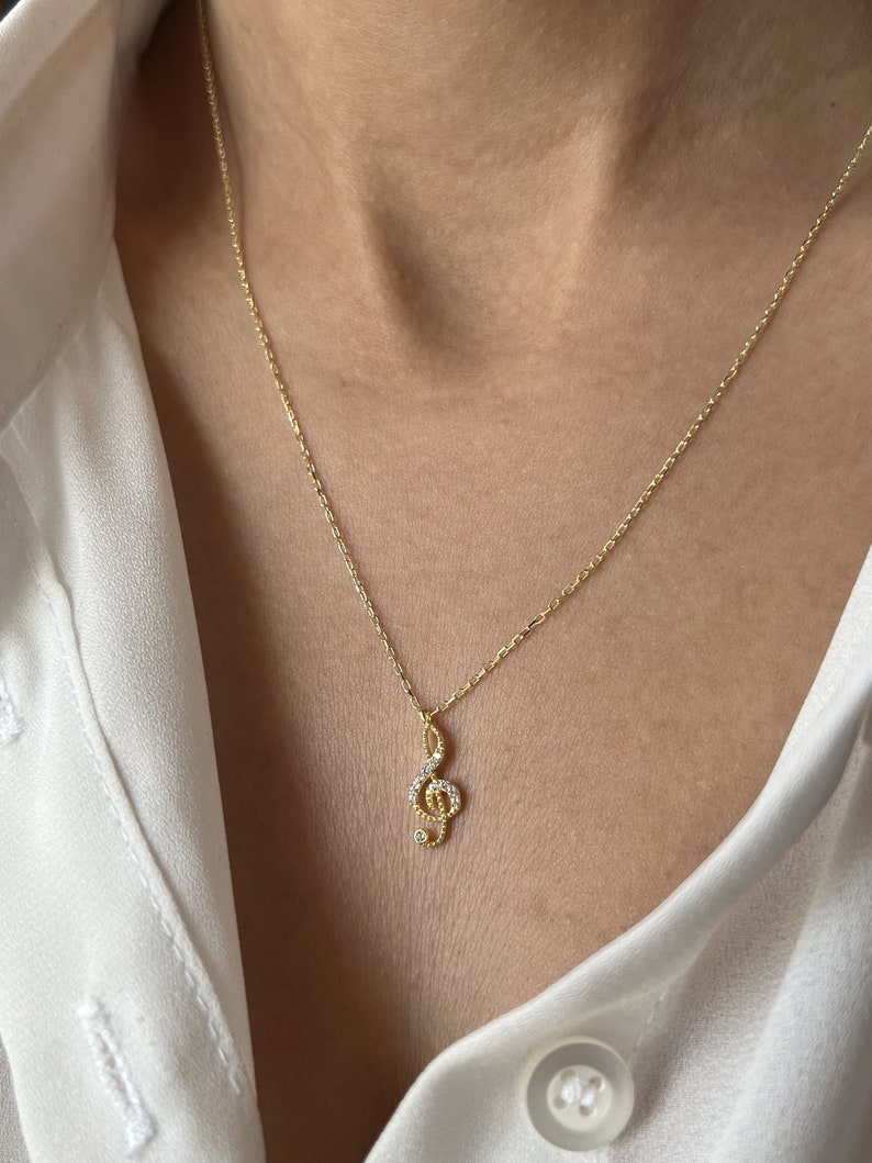 Pu&ograve; includere: Una collana con catena dorata con un piccolo ciondolo a forma di chiave di violino incrostato con pietre di zircone cubico trasparenti.