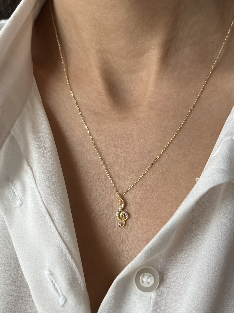 Pu&ograve; includere: Una collana con catena dorata con un piccolo ciondolo a forma di chiave di violino decorato con pietre di zircone cubico trasparenti.