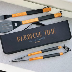 Op de afbeelding: Een barbecue gereedschapsset met een zwarte etui. De set bevat een spatel, vork en tang, allemaal met houten handvatten en zwarte accenten. Op de zwarte etui staat de tekst "BARBECUE TIME at the Smith's".