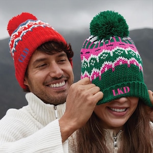 May include: A red knit beanie with a pom-pom and a blue GLD monogram, and a green knit beanie with a pom-pom, a pink, white, and green geometric pattern, and a pink LKD monogram. Both beanies feature a ribbed cuff and a thick, textured knit construction.