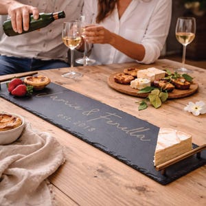 Puede incluir: Una tabla de servir de pizarra negra con asas doradas, grabada con "Jamie & Fenella est. 2018". Contiene una cuña de queso. Fresas y pasteles están sobre la mesa de madera, junto con copas de vino y una botella.