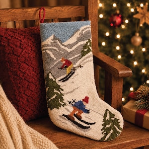 May include: A cream-colored Christmas stocking featuring a textured, hooked-wool design of a snowy mountain landscape with two skiers, green pine trees, and a red hanging loop at the top. The scene depicts skiers in colorful winter gear.