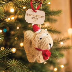 May include: A tan, textured dog ornament made of wrapped twine, wearing a small red felt Santa hat and a red scarf. Above the dog hangs a rectangular wooden tag with the engraved text MERRY CHRISTMAS OLIVER XX, suspended by a red ribbon.