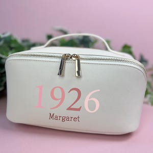 May include: A white rectangular cosmetic bag with a top handle and a gold-toned double zipper closure. The front features the pink text 1926 and the name Margaret printed in a dark pink serif font. The bag has a textured finish and rounded edges.