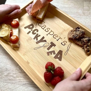 Può includere: Un vassoio rettangolare in bambù con la scritta "Casper's Picky Tea" incisa in marrone scuro. Il vassoio contiene vari snack, tra cui panini, patatine, pomodori, biscotti, fragole e prosciutto.