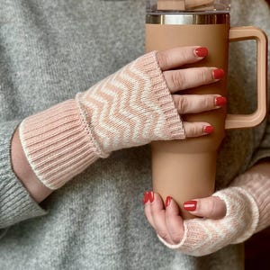 Könnte beinhalten: Ein Paar rosa-weiß gestrickte Fingerhandschuhe mit Zickzackmuster. Die Handschuhe werden getragen, und die Person hält einen großen, beigefarbenen isolierten Becher mit Griff. Der Becher hat einen transparenten Deckel.