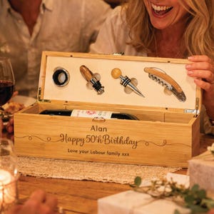May include: A wooden wine gift box containing a bottle of wine and wine accessories. The box is engraved with "Alan Happy 50th Birthday" and includes a corkscrew and bottle stoppers. The box is made of light-coloured wood.