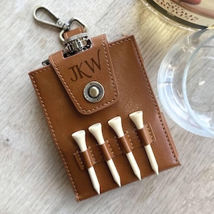 May include: A brown leather golf tee holder with a silver clasp and the initials "JKW" engraved on the flap. Four white golf tees are secured in brown leather loops. The holder is designed to attach to a golf bag.