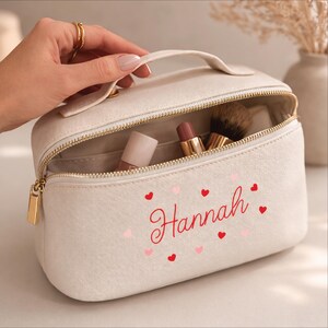 May include: Off-white makeup bag with a gold zipper and handle. The bag is open, revealing makeup items inside. The name "Hannah" is embroidered in red script with pink and red hearts.