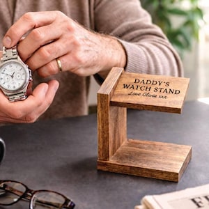 Pode incluir: Um suporte de relógio de madeira castanho escuro com o texto "DADDY'S WATCH STAND Love Olivia xxx" gravado na parte superior. O suporte tem um design de dois níveis. Um relógio prateado é segurado no fundo.