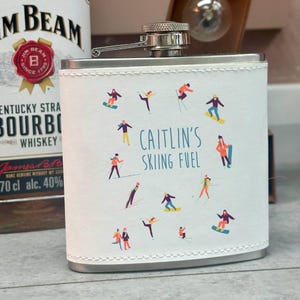 Puede incluir: Un petaca de acero inoxidable con una envoltura de cuero blanco que presenta ilustraciones de esquiadores y snowboarders. El texto "CAITLIN'S SKIING FUEL" está impreso en azul. La petaca tiene una tapa y bisagra plateadas.